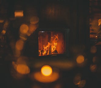 A fireplace with a crackling fire in a dark, timber/walled room. Only the fireplace in the centre of the picture is in focus. Around it are blurred golden lights that look like reflections of the fire in glas.