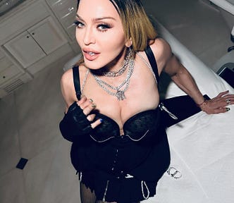 Madonna, an American iconic music performer, posing for a selfie in a mostly white tile bathroom, wearing flawless makeup, silver chains around her neck, and a black corset-esque bodice/dress and one black lace glove.
