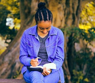 woman wearing purple jacket and jeans writing in a book while seated in front of a large tree