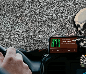 The interior of a luxury sedan as seen from the driver’s PoV, with the driver’s hands on the wheel. Mounted to the vent is a Spotify ‘Car Thing’ — a $90 tablet designed to stream Spotify music. The image has been altered. The sky behind the driver’s windscreen is animated TV static. The album thumbnail on the Car Thing UI has been replaced with a ‘code waterfall’ effect as seen in the credit sequences of the Wachowskis’ ‘Matrix’ movies. A skeletal robed figure, clutching a sheaf of documents hea