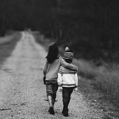 Two young children, one draping an arm over the other, both facing away from the camera, walking down a dirt/gravel road to a distance not established. Upon the sides of the road are brush that are bristlely, short, and dry grass.