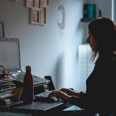 A woman sits at a cluttered desk in a dimly lit room, typing on a keyboard connected to a laptop. Her face is in profile, illuminated softly by natural light coming through a nearby window. A red water bottle, plush toy, and stacked papers fill the space around her.