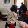 A little girl sits on a stool, facing her mother who is working at her desk, back turned to her.