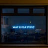 A neon sign in the window of a home office saying, “What is your story?,” with three black squares at the top of the window, and the window is surrrounded by brown bricks.