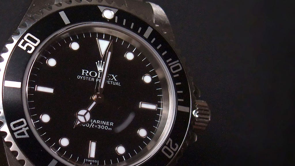 A Rolex watch With marks around the edge of the watch and dots instead of numbers on the watch face. The time is shown at just after seven. It is a silver watch with a black and white dial.