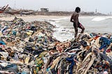 a person standing atop a heaping pile of clothes on a beach in Ghana