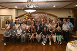 A whole lot of old people gather in a banquet room for a group photo.