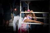 Photo of a brown-skinned child around 5–6 years old, wearing a pink mask and pink clothes and pink bow in her brown braided hair, hanging onto a bar that is probably a play structure. There’s an adult facing away behind her, maybe her parent.