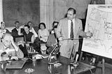 Joseph Welch (sat on the left) and Senator Joseph McCarthy (standing on the right pointing at a diagram) during the 1954 Army-McCarthy hearings. A number of people are sat in the background.