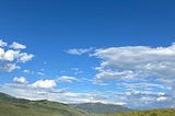 A woman walks a dirt trail within a huge field of wildflowers with a green mountain range and a blue sky dotted with clouds beyond her.