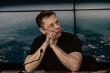 Elon Musk sits at a desk looking contemplative.