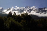 photo of snow-covered alps jutting up above some low clouds, with green trees and a tiny road sign in the foreground