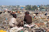 An photograph of two boys sitting on a dumpsite with grazing cows and the city in the background.