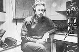 A picture from what appears to be the early 20th century of a man sitting at a desk with headphones on listening to a ham radio.