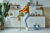 Elderly woman joyfully dances with a mop in her home kitchen.