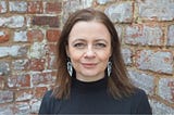 A woman with brown hair, wearing a black turtleneck, against a background of bricks.