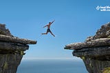Person jumping between rock ledges with blue sky and ocean in background