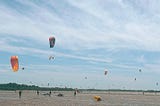 A beach on a sunny day. The sky is blue and there are wispy white clouds. On the sand there are many kite surfers, sails in the air.