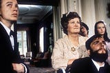 A still from the film “My Left Foot.” A man in a suit stands on the left side while a woman leans on the wheelchair of the main character, Christy Brown, played by Daniel Day-Lewis.