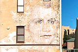 Street view of a giant, fading portrait of a woman’s face on a hotel wall. Pale yellow brickwork surrounds it.