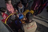 A photo shows the back of man wearing a vest that says MECA, Middle East Children’s Alliance, as he fills a water jug held by one child whilethree other children watch. Several more jugs are with the line of people waiting for water in the background.