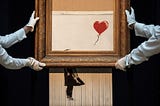 Photo of Banksy’s painting “Girl with Balloon” after it’s been shredded. It’s carried by 2 Sotheby’s employees, taking care of the newly created piece of art called “Love Is in the Bin”, carrying the art-piece with white gloves and showcasing it.