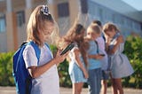 A young girl reads her cellphone alone as a group of children crowd around another cellphone behind her.