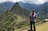 Nicole standing with blue cap, gray shirt, black pants. Background with green mountains and Machu Picchu.