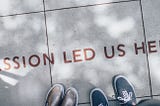 Looking down at two shoes on sun dappled concrete with the words “Passion led us here”.