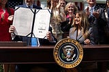 A picture showing President Donald Trump and First Lady Melania Trump participating in a bill signing ceremony for the Take it Down Act, Monday, May 19, 2025. Donald Trump is holding the bill showing their signautres while Melania is stting next to him, smiling.