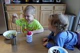 a four year old boy is eating breakfast at a talbe while his sister, 18 months old, watches