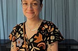 Neely stands inside an exhibit at the Blanton Museum in Austin, TX, smirking and looking to the right. She has several visible tattoos and is wearing circular glasses with orange/blue frames, dangly chain earrings, a flat gold necklace, and a short-sleeved black jumpsuit with a colorful shape pattern all over.