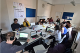 13 people sit around a long table working on laptops. Some have headphones on. A poster on the wall in the background reads ‘Design for everyone’.