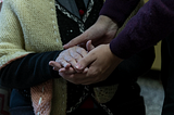 A pair of hands gently holds an elderly person’s hands wrapped in a cozy, knitted cardigan.
