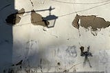 A photo of a cement wall, white paint peeling, graffiti barely covered with another coat of mismatched white. Morning sun shines on the wall, creating the shadow of a street lamp in the upper left corner.