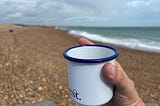 A hand holding a white cup with a blue rim on a pebble beach beside a calm sea.