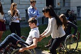 Young man in the wheelchair is looking a little scared as he tilts back in the wheelchair. His team mate is behind him to help 'spot' him. She looks equally anxious.