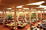A large workspace with multiple desks and workstations with high ‘lily-pad’ shaped columns and a glass ceiling, allowing for natural lighting