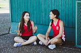 Two girls sit outside in red sports jerseys on a school playground.