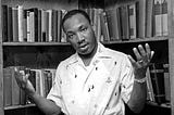 Young MLK, Jr. standing in front of a bookshelf with both of his arms raised. He has a serious look on his face.