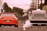 La imagen muestra una escena de la película "Fast and Furious 1" donde dos coches están en una carrera callejera. A la izquierda, hay un Toyota Supra naranja con un alerón grande y detalles de pintura verde. A la derecha, hay un Dodge Charger negro con humo saliendo de las llantas traseras, indicando que está acelerando rápidamente. Ambos coches están en una calle urbana con líneas eléctricas y árboles en el fondo.