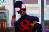 Spider-man Miles Morales stands in a black spider suit with a red spider logo on the front with the mouth part of his mask pulled up holding what appears to be a burrito maybe? Background is a bodega, which he is inside.