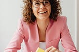 Woman in an office wearing glasses and a pink blazer holding sticky notes while looking up with a big smile on her face