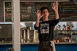 Andrew Garfield stars as Jonathan Larson in the film tick tick boom in a photo where he stands in a coffee shop dressed as a waiter holding up his hands during a musical number