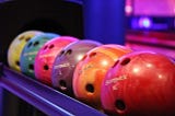 A row of colorful bowling balls labeled with various sizes (S, M, L, and XL) are displayed on a rack in a bowling alley with purple and blue lighting.