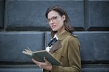 Serious adult woman wearing glasses in a smart shirt and necktie in a formal coat with open book on street against facade of old building in university. She’s looking inquisitively at the camera
