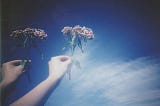 A hand holds out a small bouquet of pink flowers against a hazy blue and white background. It looks like the sky, or maybe water.