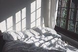 Sunlight streaming in the window, casting shadows on an unmade bed with white sheets.