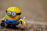 Close-up of Stewart the Minion figurine, a little smiling, bespectacled, yellow balding fellow with blue overalls, holding a banana against an outdoor dirt background.