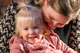 Blonde mother hugging an adorable smiling blonde toddler who is sitting on her lap.